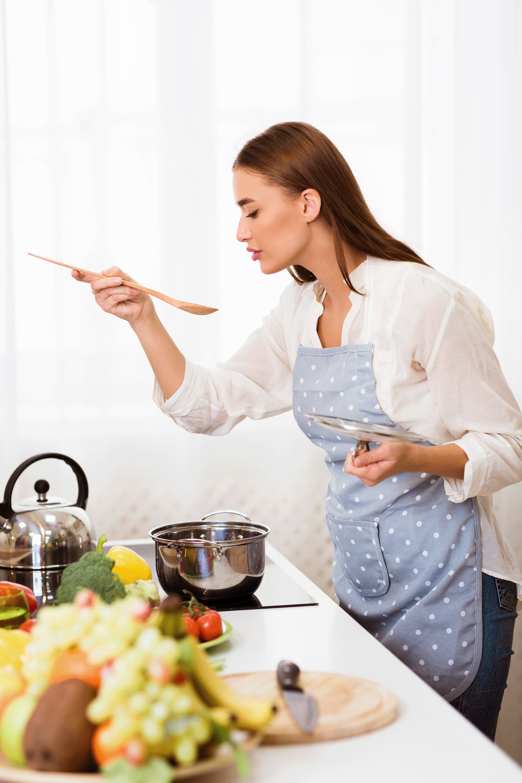 Kate Connolly carefully tasting food from a wooden spoon, checking the flavor of a dish she is cooking on the stove.