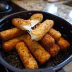 A basket of golden brown, crispy air fryer mozzarella sticks with one stick broken open showing melted, gooey cheese.