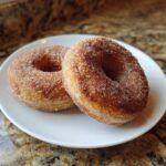 Close-up of two freshly baked apple cider donuts coated in cinnamon sugar, resting on a white plate.