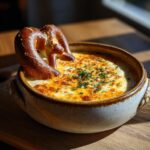 A close-up of a warm beer cheese pretzel dip topped with melted cheese and chives, with a pretzel sticking out.