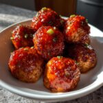 A bowl of glistening buffalo chicken meatballs, coated in a rich sauce and topped with chopped green onions.