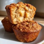 Close-up of two moist cinnamon apple muffins stacked on a white plate, one cut in half to show apple chunks.