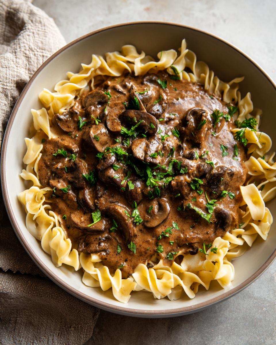 A close-up of a bowl filled with creamy mushroom stroganoff served over wide egg noodles, garnished with fresh parsley.
