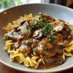A close-up of a bowl of creamy mushroom stroganoff served over egg noodles and garnished with fresh parsley.