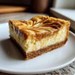 A close-up of a decadent dulce de leche cheesecake bar slice on a white plate, showcasing the swirled topping and graham cracker crust.