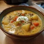A close-up of a bowl of easy cheeseburger soup, featuring ground beef, potatoes, carrots, and a dollop of sour cream with chives.