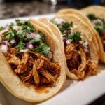 Close-up of three easy crockpot chicken tacos filled with shredded chicken and topped with diced red onion and cilantro.