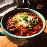 A close-up of a bowl of hearty game day chili topped with sour cream, shredded cheese, and chopped green onions.