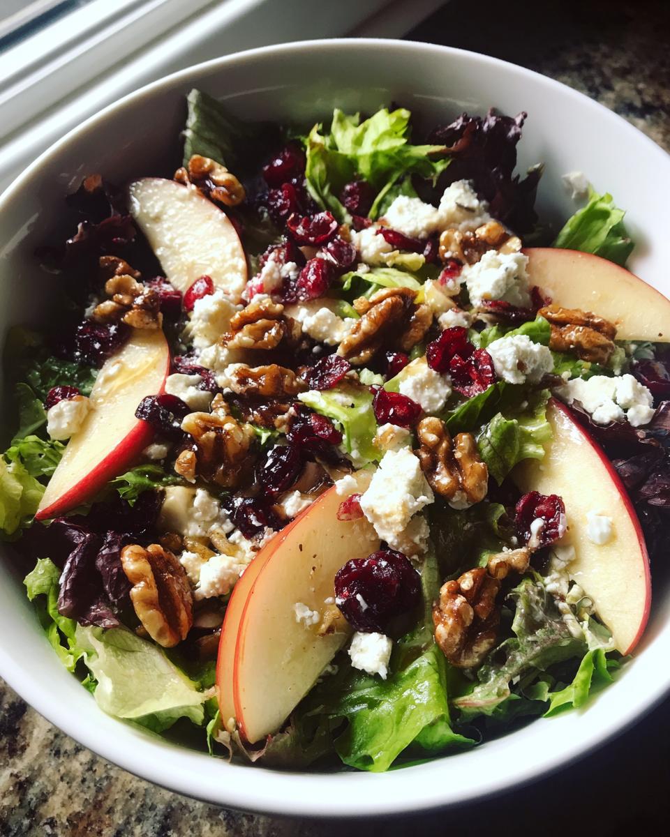 A close-up of a Harvest Apple Walnut Salad featuring fresh greens, apple slices, walnuts, feta cheese, and dried cranberries.