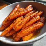 A close-up of glistening honey glazed carrots in a white bowl, seasoned with pepper.