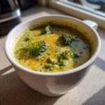 A close-up of a warm bowl of Panera broccoli cheddar soup, topped with shredded cheddar cheese and fresh broccoli florets.