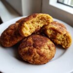 A close-up of several chewy pumpkin snickerdoodle cookies on a white plate, with one cookie broken in half.