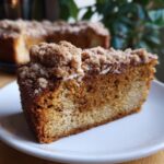 A close-up of a moist pumpkin spice coffee cake slice on a white plate, topped with crumb topping.