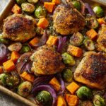 A close-up overhead view of perfectly roasted chicken thighs with Brussels sprouts, butternut squash, and red onion on a baking sheet.