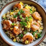 A close-up of a bowl of shrimp fried rice, featuring plump shrimp, scrambled eggs, peas, and green onions.