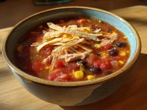 A close-up view of a bowl of slow cooker chicken tortilla soup, filled with chicken, beans, corn, and topped with tortilla strips.