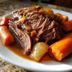 A close-up of a plate filled with shredded slow cooker pot roast, tender carrots, and soft onions in a rich gravy.