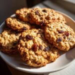 A close-up of several soft and chewy oatmeal raisin cookies piled on a white plate.