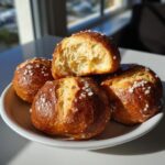 A close-up of fluffy soft pretzel bites, one broken open to reveal its airy interior, sprinkled with coarse salt.