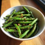 A white bowl filled with vibrant green beans tossed with spicy garlic and chili flakes.