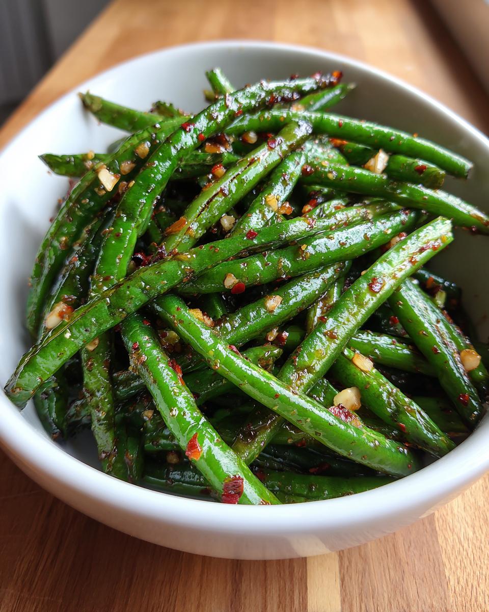 Close-up of bright green beans coated in a glossy sauce with visible garlic and chili flakes, served in a white bowl.