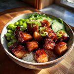 A close-up of a Teriyaki salmon bowl featuring glazed salmon cubes over rice, with edamame and sliced cucumber.