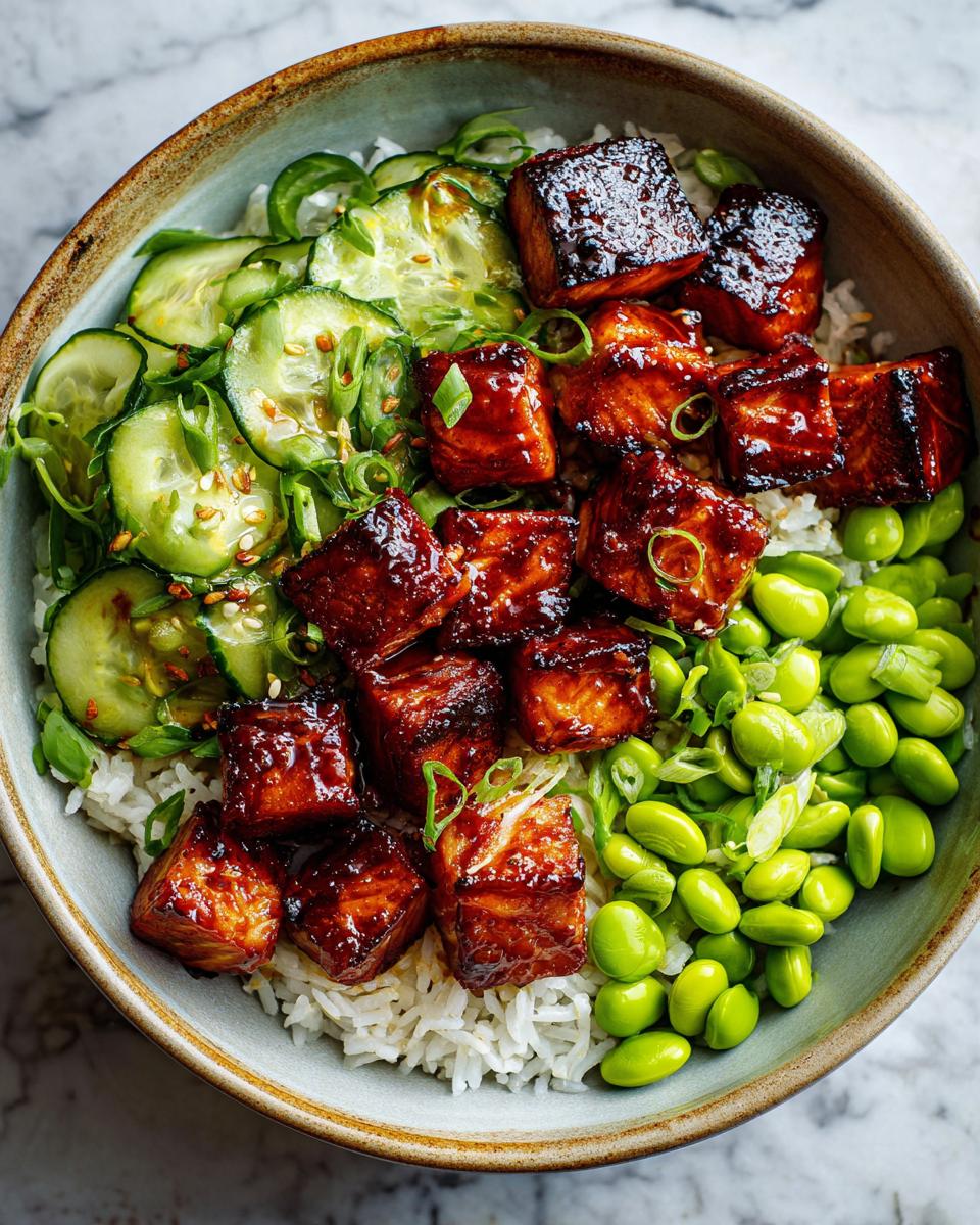A close-up of a Teriyaki Salmon Bowl featuring glazed salmon cubes, white rice, sliced cucumbers, and edamame.