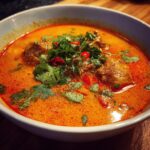 A close-up of a bowl of Thai coconut curry soup with meatballs, garnished with cilantro and chili peppers.