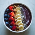 A vibrant and healthy Acai bowl topped with fresh blueberries, strawberries, banana slices, granola, chia seeds, and coconut flakes.