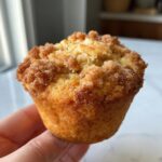 Close-up of a hand holding a perfectly baked apple cinnamon muffin with a golden-brown crust and crumbly streusel topping.