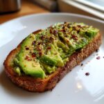 A close-up of a slice of whole-grain toast topped with mashed and sliced avocado, sprinkled with red pepper flakes and black pepper.