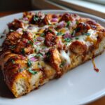 A close-up shot of a slice of BBQ chicken pizza topped with melted cheese, red onions, and parsley.
