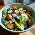 A close-up of a fresh Caesar salad in a white bowl, topped with golden croutons and grated Parmesan cheese.