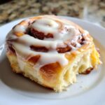 A close-up of a freshly baked cinnamon roll, generously topped with creamy white frosting.