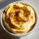 A close-up shot of creamy hummus in a white bowl, drizzled with olive oil and sprinkled with paprika and sesame seeds.