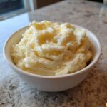 A close-up of a white bowl filled with fluffy mashed potatoes, topped with melted butter.