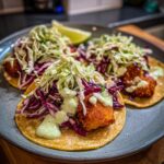 Close-up of three delicious fish tacos on a plate, topped with shredded cabbage, red cabbage, and a creamy green sauce. A lime wedge is visible.