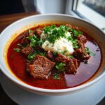 A close-up of a bowl of rich, red Goulash topped with a dollop of sour cream and fresh parsley.