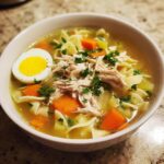 A steaming bowl of Grandma's Chicken Noodle Soup, featuring shredded chicken, wide egg noodles, carrots, celery, and a hard-boiled egg half, garnished with parsley.