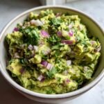 Close-up of a bowl of fresh guacamole, featuring mashed avocado, diced red onion, and chopped cilantro.