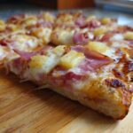 Close-up of a slice of Hawaiian pizza, showcasing melted cheese, ham, and pineapple chunks on a wooden board.