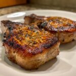 Close-up of three perfectly seared pork chops on a white plate, glistening and seasoned.
