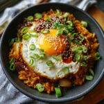 A close-up of a bowl of spicy Kimchi fried rice topped with a perfectly fried egg, sesame seeds, and chopped green onions.