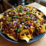 A close-up of a dark plate piled high with loaded nachos, featuring melted cheese, seasoned ground beef, black beans, and red onion.