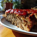 A close-up shot of a thick slice of homemade meatloaf topped with a shiny ketchup glaze, served on a white plate.
