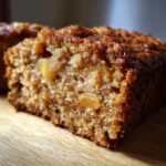 Close-up of a moist apple cake slice with visible apple pieces, on a wooden cutting board.