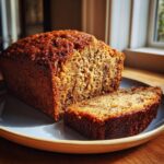 A close-up of a moist banana bread loaf with one slice cut, showcasing its texture and golden-brown crust.