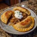 A plate of three golden-brown, pan-fried pierogi served with caramelized onions and a dollop of sour cream topped with chives.