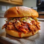 A close-up of a juicy pulled pork sandwich piled high with coleslaw and pickles on a toasted bun.