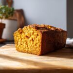 A golden-brown loaf of moist pumpkin bread on a wooden cutting board, ready to be sliced.